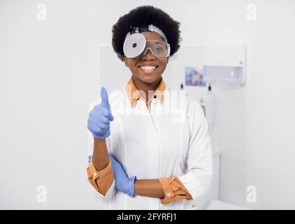 Portrait of young afro american lady otolaryngologist with a medical mirror on her head, smiling and looking at the camera, showing her thumb up. Female ENT doctor shows thumb up at modern clinic Stock Photo