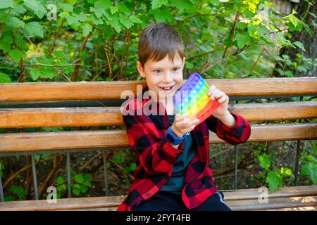 8-years old boy playing with popit in park. happy child with toy. kid wearing bright summer casual clothes. multicolored pop it close-up. Stock Photo