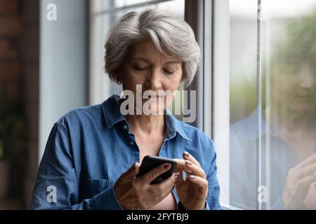 Serious mature woman standing near window with smartphone Stock Photo