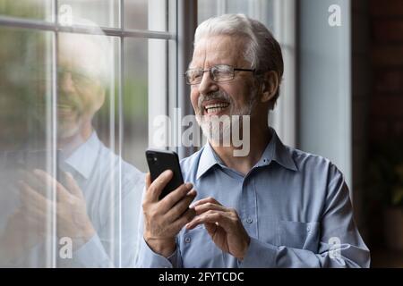 Cheerful older man holding smartphone looking out the window Stock Photo
