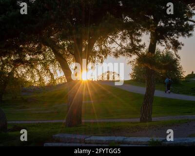 Helsinki / Finland - MAY 29, 2021: Sun casting sunrays behind a hill. A young couple riding electric scooter to the sunset. Stock Photo