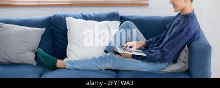 Young asian businessman smile and work from home with laptop computer online to internet on sofa in living room, freelance man using notebook on couch Stock Photo