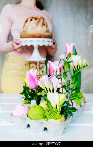 Easter floral decoration with colorful eggs, tulips, freesias and buxus. Woman holding a cake in the background. Stock Photo