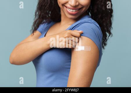 Happy covid-19 vaccinated african american woman showing arm with plaster after injection, blue background, crop Stock Photo