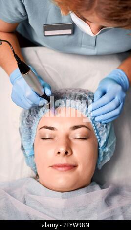 Close up of beautician hands in sterile gloves using dermapen during skincare procedure while female client closing eyes and smiling. Concept of beauty, skincare and collagen induction therapy. Stock Photo