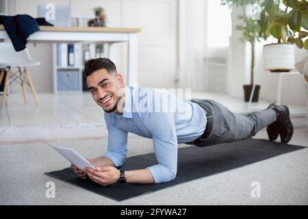 Workplace fitness. Young Arab businessman exercising on yoga mat, standing in plank and reading documents at office Stock Photo
