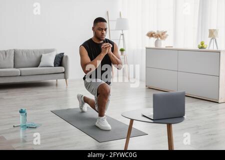 Determined African Guy Exercising At Laptop Doing Forward Lunge Indoor Stock Photo