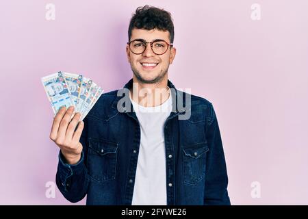 Young hispanic man holding peruvian sol banknotes looking positive and happy standing and smiling with a confident smile showing teeth Stock Photo