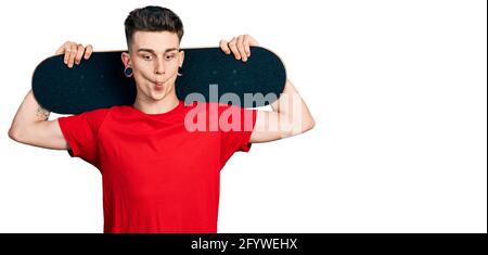 Young caucasian boy with ears dilation holding skate over shoulders making fish face with mouth and squinting eyes, crazy and comical. Stock Photo