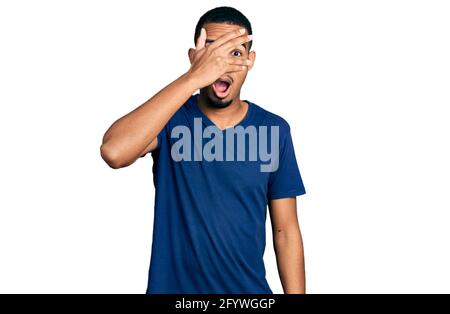 Young african american man wearing casual t shirt peeking in shock covering face and eyes with hand, looking through fingers with embarrassed expressi Stock Photo