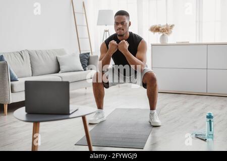 Sporty African Man Doing Deep Squats Near Laptop Computer Indoor Stock Photo