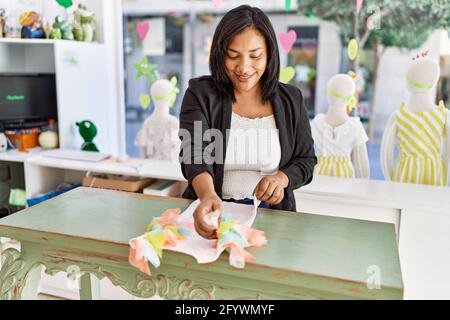Hispanic woman working as shop assistant at children clothes small retail trade. sales assistant smiling happy at shopping counter while folding shirt Stock Photo