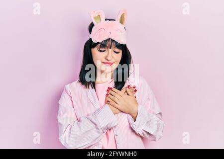 Young hispanic woman wearing sleep mask and pajama smiling with hands on chest with closed eyes and grateful gesture on face. health concept. Stock Photo