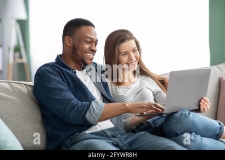 Portrait Of Happy Interracial Couple Relaxing With Laptop In Living Room Stock Photo