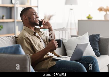 Black Man Talking On Cellphone Using Laptop Drinking Coffee Indoors Stock Photo