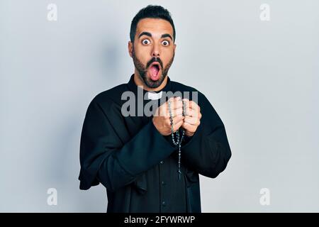Handsome hispanic priest man with beard praying holding catholic rosary afraid and shocked with surprise and amazed expression, fear and excited face. Stock Photo