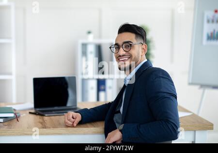 Happy Arab businessman sitting at table with laptop computer at company office, mockup for website design Stock Photo