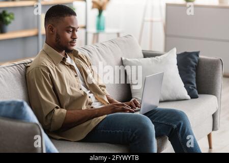 Serious Black Man Using Laptop Browsing Internet Sitting At Home Stock Photo