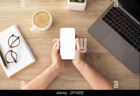 Top view of female hands holding smartphone with blank screen, laptop keyboard, cup of coffee on wooden table Stock Photo