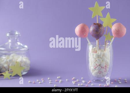 glazed cake pops in a glass and a jar with marshmallows on a purple background with sprinkles. candy bar concept. horizontal image. place for text.