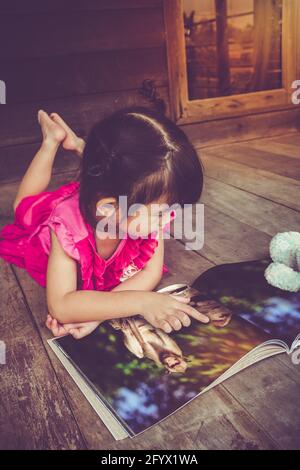 Closeup handsome asian girl smiling and reading picture album, lying on wooden floor with bright sunlight, outdoor at home. Children read and study, e Stock Photo