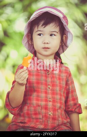 Little asian girl wearing pink hat and eating ice cream in the summer on blurred nature background. Pretty child in nature. Outdoors. Cross process. Stock Photo