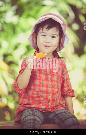 Little asian girl wearing pink hat and eating ice cream in the summer on blurred nature background. Pretty child in nature. Outdoors. Cross process. Stock Photo