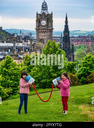 Two young girls pretend to communicate with giant connected tin cans launching Edinburgh Science Festival, Calton Hill with city skyline, Scotland, UK Stock Photo