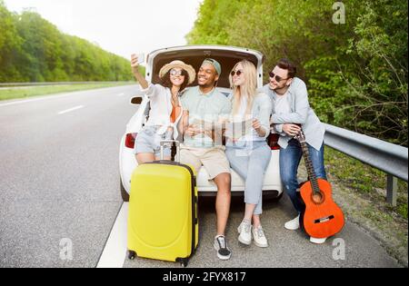 Group of best friends checking map during road trip, sitting in car trunk, taking selfie on their journey. Hitchhiking Stock Photo