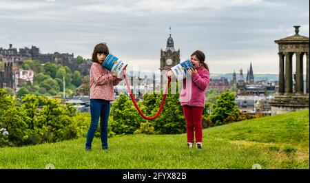 Two young girls pretend to communicate with giant connected tin cans launching Edinburgh Science Festival, Calton Hill with city skyline, Scotland, UK Stock Photo