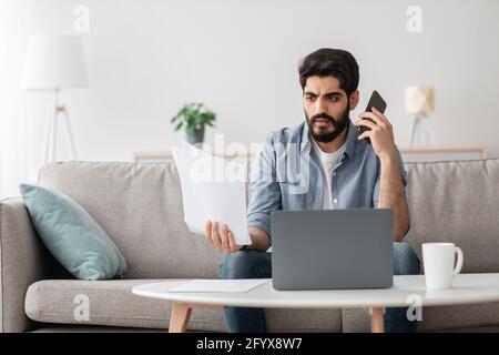 Problems at work. Arab man discussing documents, holding paper and talking on cellphone, working remotely at home Stock Photo