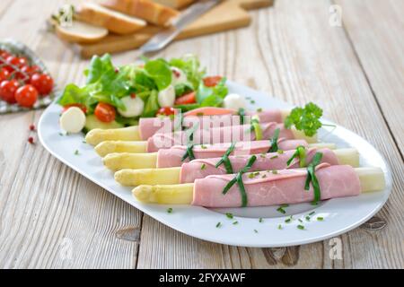 Delicious spring salad with ham asparagus rolls on a white platter, served with baguette slices on a wooden table Stock Photo
