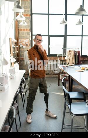 Young man with leg prosthesis standing between tables in workshop Stock Photo