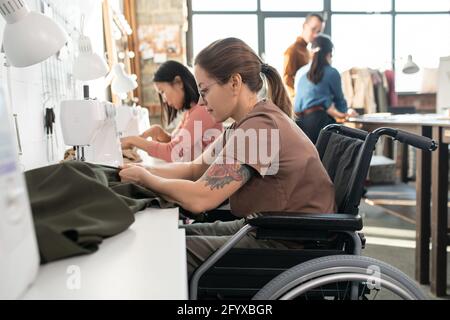 Two females sewing clothes in workshop against colleagues Stock Photo