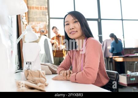 Happy young seamstress sitting by sewing machine in workshop Stock Photo