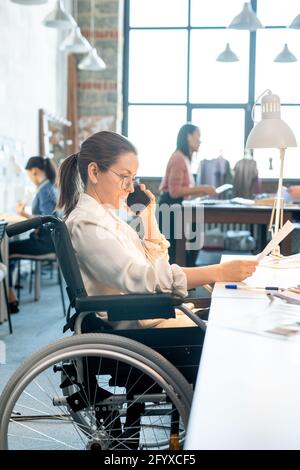 Female in wheelchair looking through paper and talking to client on smartphone Stock Photo