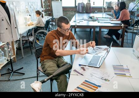 Young disable man sitting by table and working over fashion collection in large workshop Stock Photo