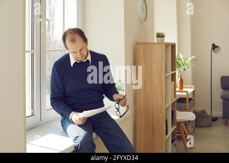 Mature businessman working with paper sitting on windowsill in home office Stock Photo