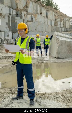 Young builder with sketch standing on construction site against two colleagues Stock Photo