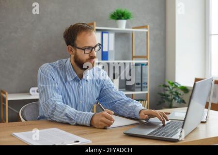 Young serious concentrated man working on laptop writing notes on paper sheet Stock Photo