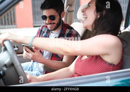 Two friends travel together driving a car, using the smartphone and laughing. Summer adventure. Stock Photo