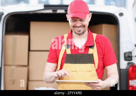 Male courier in uniform stands near car with boxes and holds tablet Stock Photo