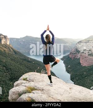 Back view of faceless fit female hiker in activewear standing in Tree with Arms Up asana while practicing yoga on rocky cliff over river in canyon Stock Photo