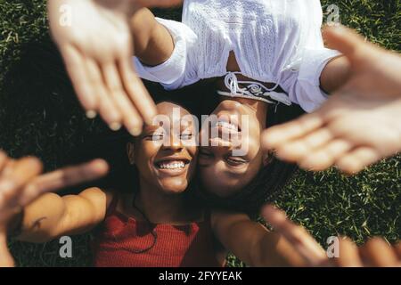 Top view of smiling African American female friends reaching hands to camera while lying on grassy lawn on sunny summer day Stock Photo