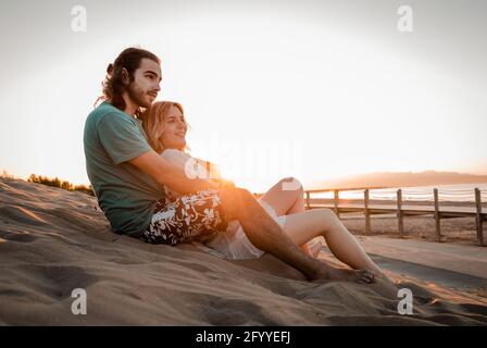 Full body side view young serene couple in summer wear hugging and lying on warm sandy beach at picturesque sunset Stock Photo