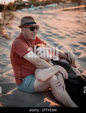 Content adult couple in summer wear embracing and resting on sandy seacoast during scenic sunset Stock Photo