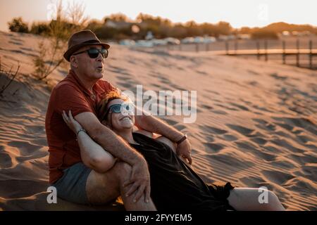 Content adult couple in summer wear embracing and resting on sandy seacoast during scenic sunset Stock Photo