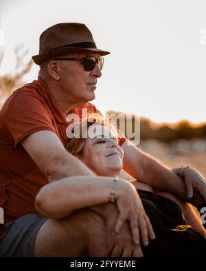 Content adult couple in summer wear embracing and resting on sandy seacoast during scenic sunset Stock Photo
