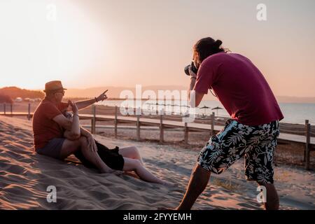 Focused male photographer taking pictures of content adult couple hugging and resting on sandy seacoast at sunset Stock Photo
