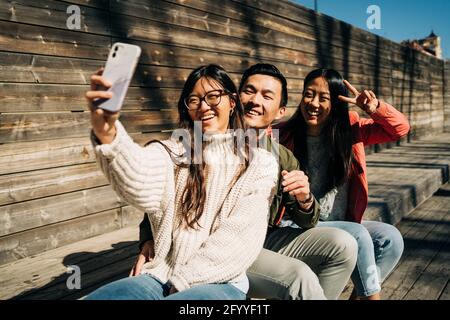 Cheerful young Asian friends in stylish outfits smiling and showing two fingers sign while taking selfie on smartphone sitting on wooden bench on sunn Stock Photo